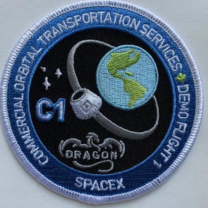 May include: Embroidered patch with a blue border and the text "COMMERCIAL ORBITAL TRANSPORTATION SERVICES DEMO FLIGHT 1." The patch features a globe, a spacecraft, and the text "C1," "DRAGON," and "SPACEX."