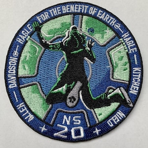 Puede incluir: Un parche bordado con fondo azul y contorno blanco. El parche presenta una silueta de una persona saltando con un diseño de mapa verde y azul dentro del círculo. El texto alrededor del círculo dice "ALLEN - DAVIDSON - HAGLE FOR THE BENEFIT OF EARTH - HAGLE - KITCHEN" y "NS 20".