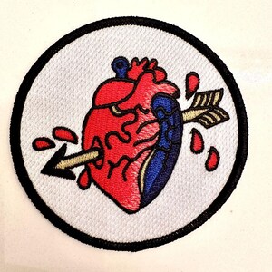 May include: Embroidered patch featuring a stylized anatomical heart pierced by a gold arrow. The heart is red and blue, with red droplets and a black border on a white background. A classic tattoo-inspired design.