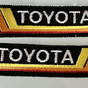 Puede incluir: Dos parches de tela negra con la palabra "TOYOTA" bordada en blanco. Cada parche tiene un diseño de rayas rojas, naranjas y amarillas.