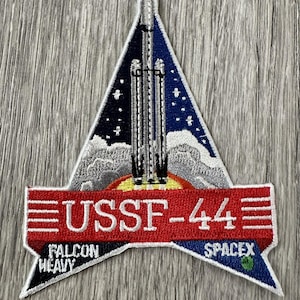 May include: A triangular patch with a red, white, and blue design. It features a rocket launching into space with a white cloud and a yellow sun. The text "USSF-44" is in white on a red banner. The words "FALCON HEAVY" and "SPACEX" are on the bottom corners of the patch.