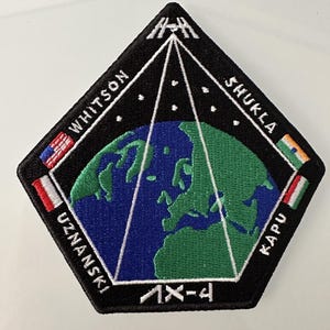 Może przedstawiać: Naszywka haftowana w kolorze czarnym i białym z przedstawieniem stacji kosmicznej, globu ziemskiego oraz nazwiskami "Whitson", "Shukla", "Uznanski" i "Karp" wokół globu. Na dole naszywki widnieje napis "AX-4".