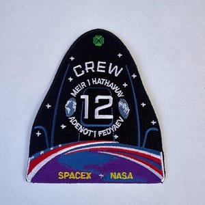 Peut inclure: Un écusson brodé noir en forme de dôme avec le texte CREW 12, MEIR, HATHAWAY, ADENOT et FEDYAEV. Il présente un trèfle à quatre feuilles vert, des étoiles blanches, une Terre bleue et des arcs rouge, blanc et bleu. SPACEX et NASA sont inscrits en bas.