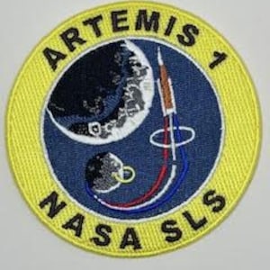 May include: A round, yellow patch with the text "ARTEMIS 1 NASA SLS" around the edge. The center of the patch features a blue background with a white moon and a rocket launching from the moon's surface.