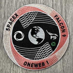 May include: Embroidered patch with the text "SPACEX FALCON 9 ONEWEB 1". The patch features a black center with a white Earth, a stylized Falcon 9 rocket, and a white and pink outer ring with a net-like design.