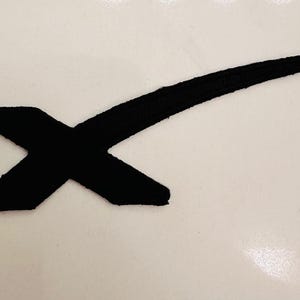 Può includere: Toppa in feltro nero con una X stilizzata e una linea curva che si estende dall'angolo in alto a destra.