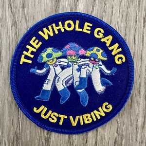Puede incluir: Un parche redondo azul con texto amarillo que dice "THE WHOLE GANG JUST VIBING". El parche presenta tres figuras con forma de hongo con trajes espaciales blancos y gorras coloridas. Probablemente un parche para planchar o coser.