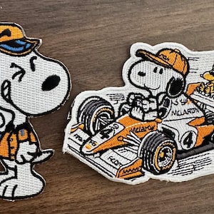 Op de afbeelding: Geborduurde patches met Snoopy in een oranje en witte race-outfit en Snoopy in een raceauto. De patches hebben zwarte contouren en liggen op een houten oppervlak. Op de raceauto staat het woord "McLaren".