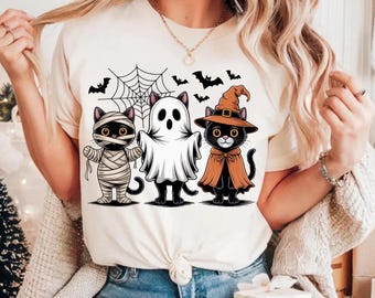 May include: A cream-colored t-shirt featuring a Halloween-themed graphic. The design includes three cartoon cats dressed as a mummy, a ghost, and a witch, with bats and a spiderweb above them. The cats are black with yellow eyes.