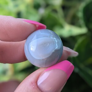 May include: A smooth, round, grey agate stone with a white centre. The stone is being held between two fingers.