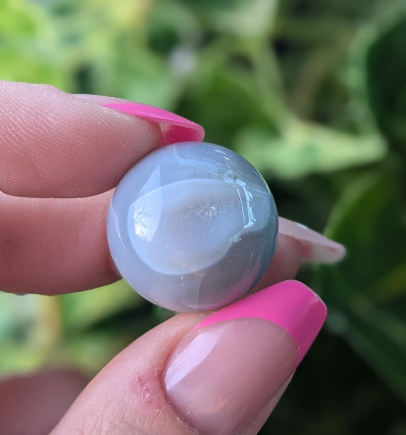 May include: A smooth, round, grey agate stone with a white centre. The stone is being held between two fingers.