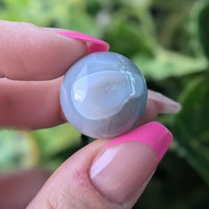 May include: A smooth, round, grey agate sphere with a white centre. The sphere is being held in a hand with pink nail polish.