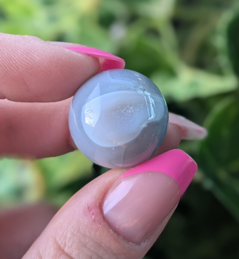 May include: A smooth, round, grey agate sphere with a white centre. The sphere is being held in a hand with pink nail polish.