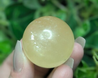 May include: A smooth, pale yellow, polished sphere of calcite crystal. The sphere is being held in a hand.