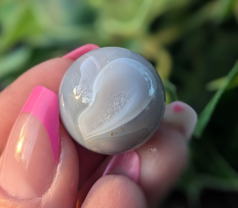 May include: A smooth, grey agate sphere with white swirling patterns. The sphere is about the size of a marble.