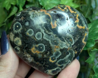 May include: A heart-shaped polished stone with a black, green, and orange pattern. The stone has a unique, swirling pattern with small, round, white spots.