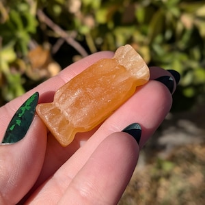 Puede incluir: Una piedra translúcida en forma de caramelo de color ámbar. La piedra se sostiene en una mano, con un fondo borroso verde y marrón. La piedra en forma de caramelo tiene la apariencia de un caramelo envuelto.