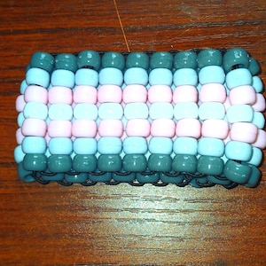 May include: A handmade beaded bracelet featuring a pattern of gray, light blue, and pink beads. The bracelet is made of small, round beads strung together, creating a colorful accessory.