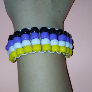 May include: A beaded bracelet with a non-binary pride flag design. The bracelet features rows of yellow, white, purple, and black beads strung together with white string. The bracelet is worn on a wrist.