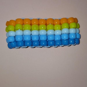 May include: A handmade bracelet composed of colorful plastic beads. The bracelet features rows of orange, yellow, green, and blue beads, creating a vibrant, layered effect. The beads are strung together with a white elastic cord.