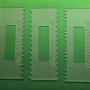 May include: Three clear plastic cake scrapers with different edge designs: scalloped, zig-zag, and straight. Each scraper has a rectangular cutout and the text "The Velvet Whisk" in a script font. The background is a solid green.
