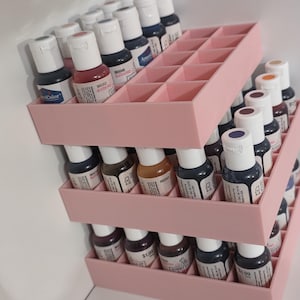 May include: A three-tiered light pink plastic storage rack with square compartments holding multiple small bottles of craft paint. Each bottle features a white cap and a label with printed text, organized neatly within the grid-style shelves.