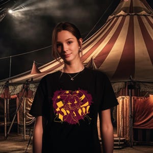 May include: Black t-shirt featuring a graphic design with a stylized face, stars, and stripes in yellow and burgundy. The design is set against a dark background, with a circus tent visible in the background.