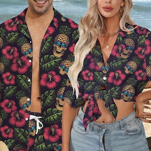 May include: A couple wearing matching black and green Hawaiian shirts with a skull and pineapple print. The shirts have red hibiscus flowers and green leaves. The woman's shirt is tied at the waist.