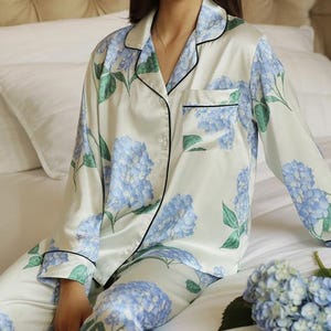 Può includere: Completo pigiama bianco con stampa floreale di ortensie blu. La parte superiore a maniche lunghe e i pantaloni hanno bordi neri. Un bouquet di ortensie blu è appoggiato sul letto. Il pigiama è in materiale setoso.