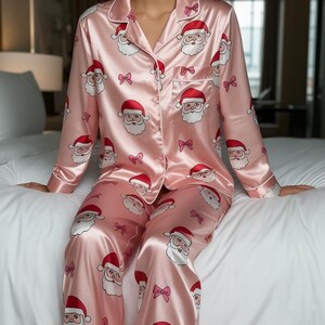 Könnte beinhalten: Hellrosa Satin-Pyjama-Set mit einem Muster aus Weihnachtsmann-Gesichtern und Schleifen. Das langärmlige Oberteil hat eine Knopfleiste und die Hose hat eine lockere Passform. Der Pyjama ist aus glänzendem, seidigem Material gefertigt.