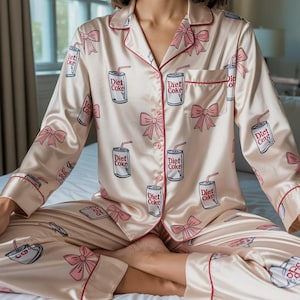 Op de afbeelding: Lichtroze satijnen pyjamaset met een herhalend patroon van Diet Coke-blikjes en roze strikken. De set bevat een overhemd met knopen en rode biezen en een bijpassende broek. De pyjama's zijn ontworpen voor comfort en stijl.