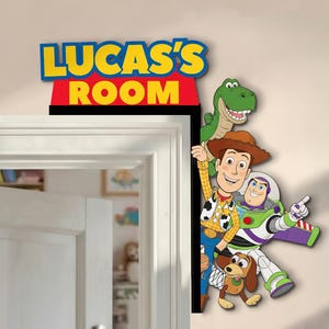 Op de afbeelding: Kleurrijke wanddecoratie met personages uit de film Toy Story. Het bord erboven leest "LUCAS'S ROOM" in geel en rood. De personages zijn Woody, Buzz Lightyear, Rex en Slinky Dog. Geschikt voor een kinderkamer.