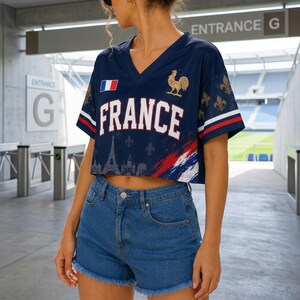 May include: A navy blue V-neck cropped t-shirt featuring the word FRANCE in white letters, a French flag, a gold rooster emblem, and a graphic of the Eiffel Tower. The sleeves have red and white stripes and gold fleur-de-lis patterns.