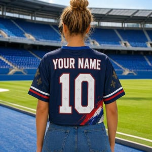 May include: A dark blue short-sleeved jersey featuring the text YOUR NAME and the number 10 in white with red outlines. The sleeves have gold fleur-de-lis patterns and red and white stripes, with a red, white, and blue brushstroke design.