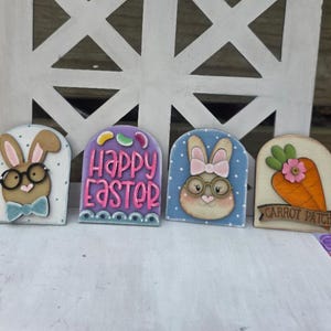 May include: Four wooden Easter-themed decorations. One features a bunny with glasses and a bow tie, another says "Happy Easter", a third shows a bunny with a pink bow, and the last has a carrot with a flower and the words "Carrot Patch".