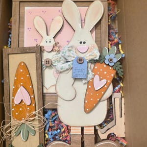 May include: Wooden Easter decorations featuring a large bunny holding a carrot with a pink heart, and a smaller bunny. The large bunny has a tag that says "Fresh Carrots". The decorations are in a cardboard box with colorful paper filler.