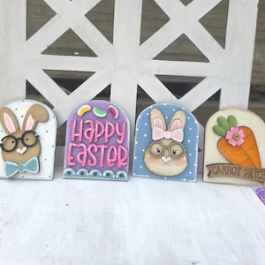 May include: Four Easter-themed wooden decorations. One features a bunny with glasses and a bow tie, another says "Happy Easter", and a third shows a bunny with a pink bow. The last one has a carrot with a flower and the words "Carrot Patch".