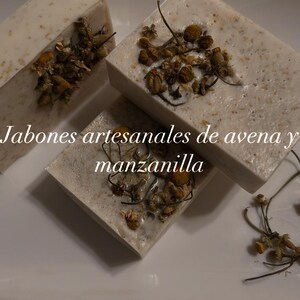 May include: Three white bars of soap with dried chamomile flowers and the text "Jabones artesanales de avena y manzanilla" on top of the bars.