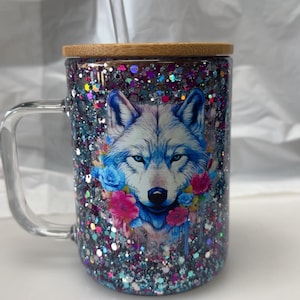 May include: A clear glass mug with a handle, filled with glitter and a wolf design. The wolf has blue eyes and is surrounded by flowers. It includes a bamboo lid and a clear straw.
