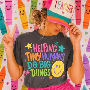 May include: A charcoal gray t-shirt featuring the colorful text Helping Tiny Humans Do Big Things with a yellow smiley face and stars, paired with a white and teal trucker hat with the word Teacher printed in pink on the front panel.