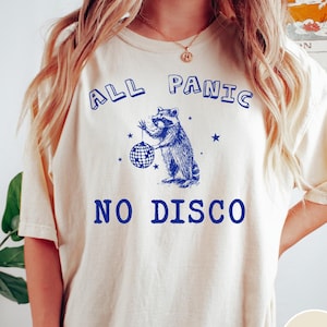 May include: A cream coloured t-shirt with blue text that reads "All Panic No Disco". A cartoon raccoon holding a disco ball is in the centre of the text. The word "Ivory" is in a brown rectangle in the bottom right corner of the image.