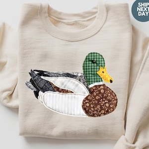 May include: A cream-coloured sweatshirt with a duck appliqué. The duck has a green and white checked head, yellow beak, and a brown floral patterned body. The wings are a mix of grey, white, and brown fabrics. The text "SHIP NEXT DAY" is in a circle.