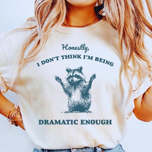 May include: A white t-shirt with a blue raccoon graphic and the text "Honestly, I don't think I'm being dramatic enough".