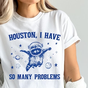 Può includere: Maglietta bianca con testo blu che recita "Houston, ho tanti problemi". Un cartone animato di un procione con un casco da astronauta che galleggia nello spazio con pianeti e stelle.