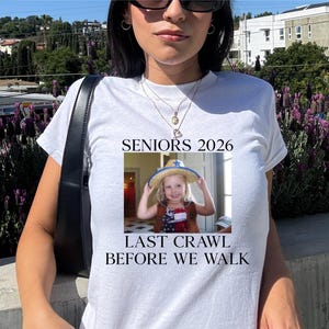 May include: White t-shirt with the text "SENIORS 2026" and a photo of a young child wearing a hat. The text "LAST CRAWL BEFORE WE WALK" is below the photo. The shirt is being worn by a person.
