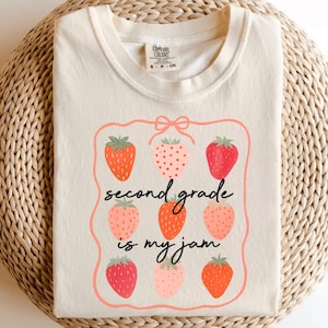 Könnte beinhalten: Elfenbeinfarbenes T-Shirt mit Rundhalsausschnitt. Das Shirt zeigt eine Erdbeer-Grafik mit dem Text "second grade is my jam" in Schwarz. Die Erdbeeren sind in Rot- und Rosatönen gehalten.