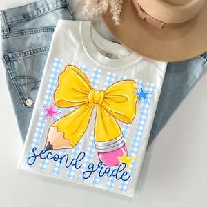 Può includere: Una t-shirt bianca con la grafica di una matita gialla a forma di fiocco, su uno sfondo a quadretti vichy azzurro. Il design include piccole stelle rosa, blu e gialle e il testo in corsivo blu second grade.