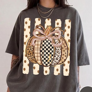 Może przedstawiać: Ciemnoszary t-shirt z krótkim rękawem z centralną grafiką dyni z jasnoróżową kokardą. Dynia posiada sekcje z motywem panterki, kraty i szachownicy, umieszczone na tle pionowych beżowych pasów z brązowymi kropkami.
