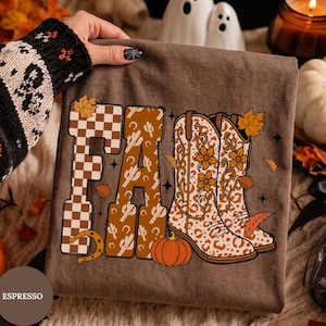 Könnte beinhalten: Ein espressofarbenes T-Shirt mit dem Wort FALL in großen Buchstaben. Das F ist ein braun-weißes Schachbrettmuster, das A hat ein Kaktusmuster und die LL sind zwei Cowboystiefel mit Blumen- und Leopardenmuster. Enthält Kürbisse und Blätter.