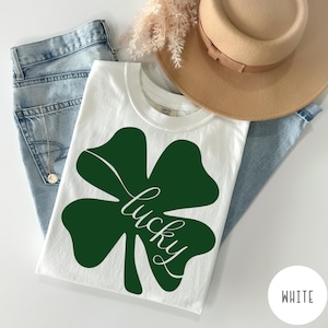 Puede incluir: Una camiseta blanca con un gran diseño de trébol verde y la palabra "lucky" en cursiva. La camiseta se combina con vaqueros azul claro, un sombrero beige y flores secas. Un círculo blanco con la palabra "WHITE" está en la esquina inferior derecha.